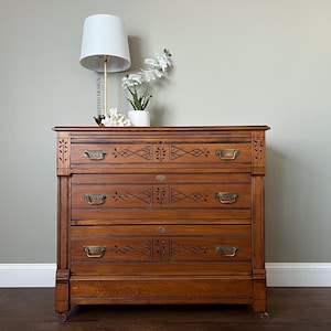 May include: A vintage wooden dresser with three drawers and intricate carved details. The dresser has brass hardware and is painted in a warm brown color.
