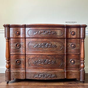 May include: Antique wooden dresser with three drawers. The dresser is a dark brown color with ornate carvings on the drawer fronts and round knobs. The sides have decorative columns. The dresser is a classic piece of furniture.