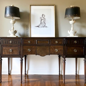 May include: A dark wood sideboard with four drawers and six legs. Two lamps with black and gold lampshades are on top of the sideboard. A framed print of a woman sitting with a white background is hanging on the wall behind the sideboard.