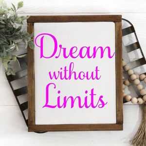 Op de afbeelding: Een houten bord met een witte achtergrond en een bruin frame. Het bord zegt "Dream without Limits" in roze letters.