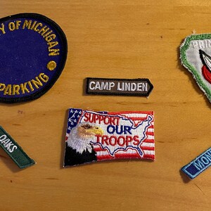May include: A collection of six embroidered patches. The patches are various shapes and sizes and feature different designs, including a university logo, a military support message, a camp name, and a princess logo. The patches are all on a wooden surface.