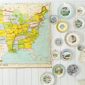 May include: A white wall with a vintage map of the United States and a collection of decorative plates featuring state-themed designs. The plates are arranged in a staggered pattern and are in various colors and sizes.