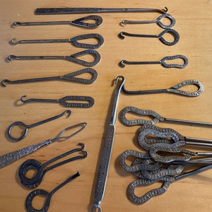 May include: A collection of vintage metal tools, including crochet hooks and other implements, arranged on a light wooden surface. Some tools have looped handles and stamped text, while others have intricate designs.