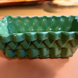 May include: A rectangular, green ceramic dish with a textured, geometric design. The dish has a scalloped rim and a series of stacked, triangular shapes forming the sides. The color is a muted, medium green.