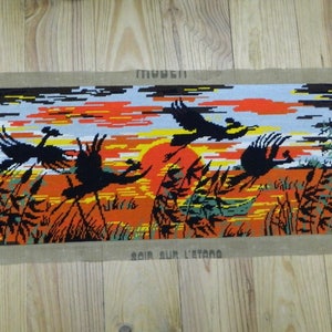 May include: Needlepoint tapestry depicting a sunset scene with flying birds over a marsh. The artwork features a vibrant orange and yellow sky, black birds, and tall grass. The words "SOIR SUR L'ETANG" are visible.