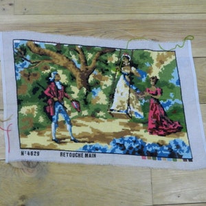 以下が含まれることがあります： 3人の人物が時代衣装を着た牧歌的なシーンを描いた、ヴィンテージのニードルポイントタペストリー。赤いコートと青いズボンを着た男性と、ドレスを着た2人の女性が特徴です。タペストリーは未完成で、糸がほつれており、「N° 4629 RETOUCHE MAIN」の文字があります。