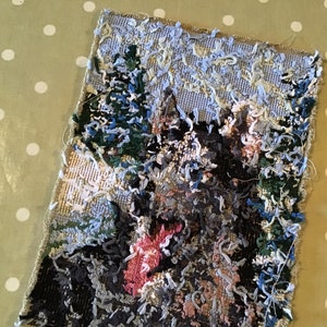 May include: A rectangular textile art piece depicting a landscape scene with trees and a mountain. The artwork features a textured, raised surface with various colors, including green, blue, pink, and gray. The edges of the textile are frayed.
