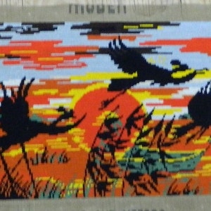 May include: Needlepoint tapestry depicting a sunset scene with silhouetted birds in flight. The artwork features a vibrant orange and yellow sky, with black birds and tall grass in the foreground. The tapestry is rectangular and mounted on a neutral-colored backing.