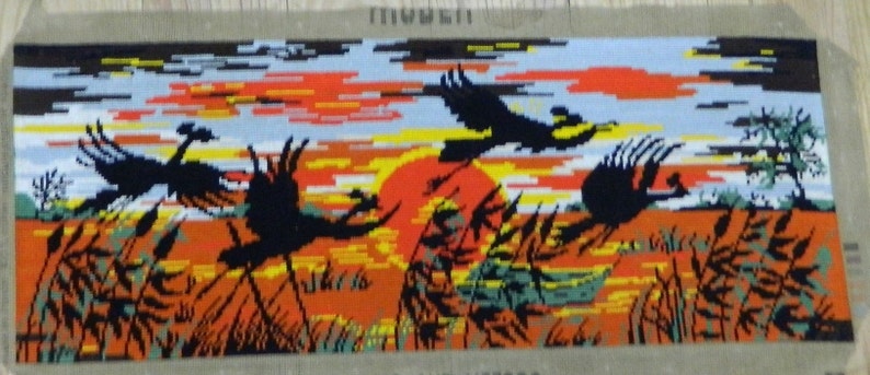 May include: Needlepoint tapestry depicting a sunset scene with silhouetted birds in flight. The artwork features a vibrant orange and yellow sky, with black birds and tall grass in the foreground. The tapestry is rectangular and mounted on a neutral-colored backing.