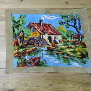 May include: A vintage needlepoint tapestry depicting a serene waterside scene. The artwork features a house with a red roof, a water wheel, and a person fishing from a boat. The tapestry is framed by a light brown border.