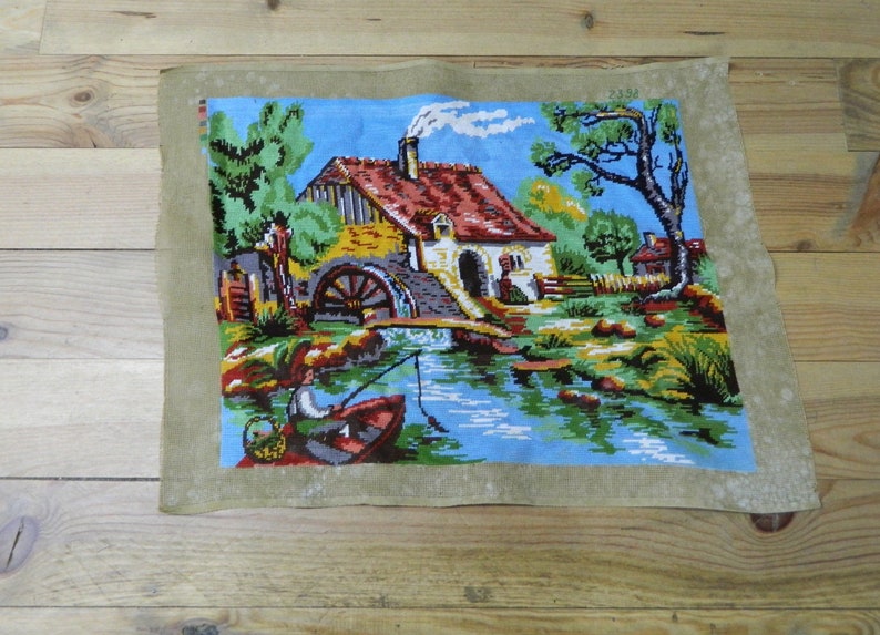 May include: A vintage needlepoint tapestry depicting a serene waterside scene. The artwork features a house with a red roof, a water wheel, and a person fishing from a boat. The tapestry is framed by a light brown border.