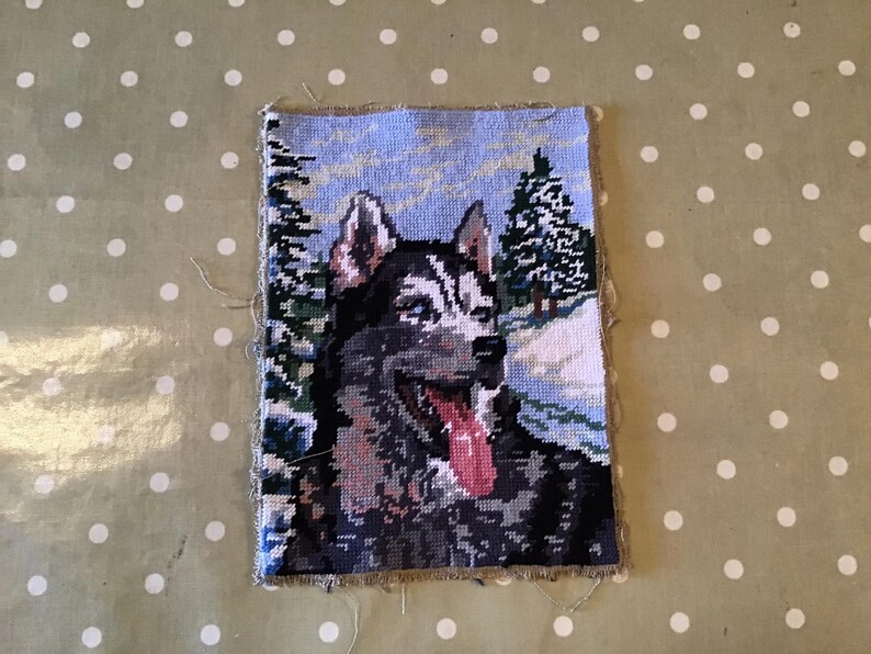 May include: A cross-stitch tapestry featuring a Siberian Husky dog in a winter landscape. The dog has black and white fur with a pink tongue. The background shows snow-covered trees and a light blue sky. The tapestry is rectangular with frayed edges.