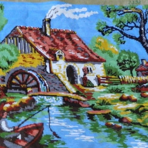 May include: A cross-stitch tapestry depicting a serene waterside scene. A red-roofed cottage with a water wheel sits beside a river, with a person fishing from a boat. The sky is blue, and the landscape is filled with trees and greenery.