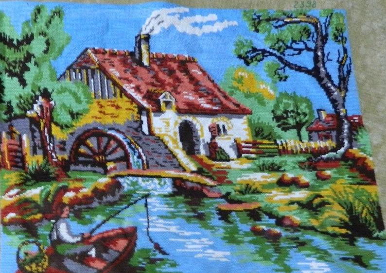 May include: A cross-stitch tapestry depicting a serene waterside scene. A red-roofed cottage with a water wheel sits beside a river, with a person fishing from a boat. The sky is blue, and the landscape is filled with trees and greenery.
