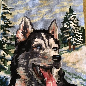 May include: Cross-stitch textile featuring a Siberian Husky dog with blue eyes, a black and white coat, and a pink tongue. The background depicts a snowy landscape with evergreen trees and a light blue sky.