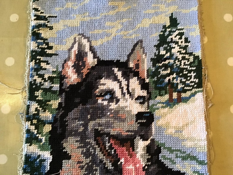 May include: Cross-stitch textile featuring a Siberian Husky dog with blue eyes, a black and white coat, and a pink tongue. The background depicts a snowy landscape with evergreen trees and a light blue sky.