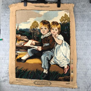 May include: A vintage tapestry featuring two children, a boy and a girl, seated together. The boy holds a drawing pad and pencil. The artwork depicts a landscape with trees, a church, and a cloudy sky. The tapestry is framed with a beige border.