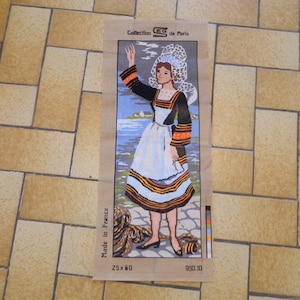 May include: Needlepoint tapestry featuring a woman in traditional Breton attire, with a white lace headdress and apron, black sleeves with orange accents, and a blue and white skirt. The tapestry is labeled "Collection de Paris" and measures 25 x 60.