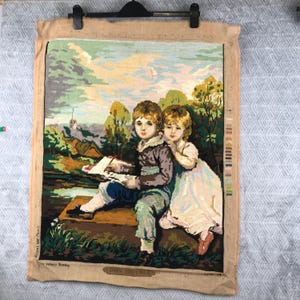 May include: A tapestry depicting two children in a landscape scene. The boy is holding a book, and the girl is leaning on his shoulder. The artwork features a variety of colors, including green, blue, brown, and beige. The tapestry is framed and ready to hang.