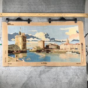 Puede incluir: Un tapiz que representa una escena costera con una torre de piedra, barcos y edificios bajo un cielo azul. El tapiz mide 165 x 51 cm. El texto "La Rochelle" es visible.