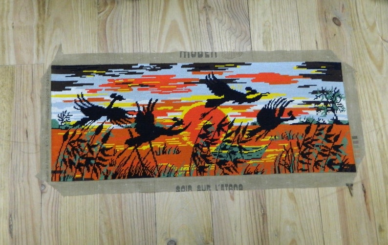 May include: Needlepoint tapestry depicting a sunset scene with flying birds over a marsh. The artwork features a vibrant orange and yellow sky, black birds, and tall grass. The words "SOIR SUR L'ETANG" are visible.