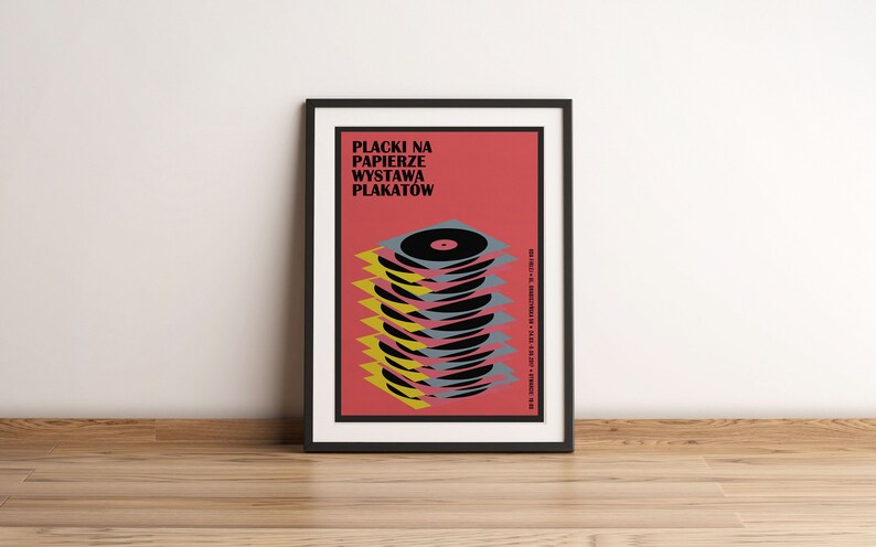 May include: A black framed poster with a red background and the text "Placki na Papierze Wystawa Plakatow". The poster features a stack of black vinyl records with yellow and grey accents.