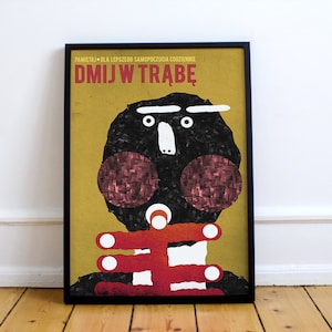 May include: A yellow and black poster with a cartoon face and the text "DMJW TRABĘ". The poster is framed in black.