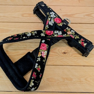 May include: Black dog harness with a floral pattern featuring pink and white roses. The harness has a padded chest strap and adjustable straps with black plastic buckles and a metal D-ring for leash attachment.