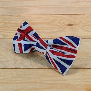 May include: A bow tie featuring the Union Jack flag design in red, white, and blue. The bow tie is made of fabric and is knotted in the center. The background is a light brown wooden surface.
