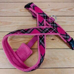May include: A pink dog harness with a plaid pattern in black and white. The harness has a soft, fuzzy lining and adjustable straps with black plastic buckles. A metal ring is attached for leash attachment.