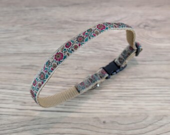 May include: A pet collar featuring a beige nylon webbing base with a decorative floral ribbon overlay. The ribbon displays a repeating pattern of pink, teal, and brown flowers with green leaves. It includes a black plastic side-release buckle.