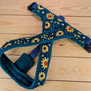 May include: A teal dog harness with a sunflower pattern. The harness features yellow sunflowers with brown centers, green leaves, and white star-shaped accents on a dark teal background. The harness has blue straps and black buckles.