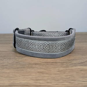 May include: A grey velvet pet collar featuring a silver metallic woven Celtic knot pattern running along the center. The collar includes black metal hardware, including a buckle and D-ring, and is constructed with a soft, plush fabric texture.