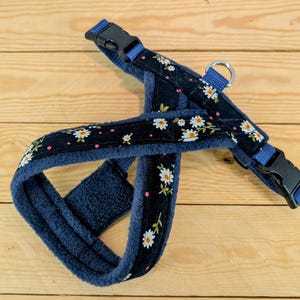 May include: A navy blue dog harness with a daisy floral pattern. The harness has a soft, blue interior and features black buckles and a metal D-ring for leash attachment. The daisy pattern includes white flowers and pink dots.