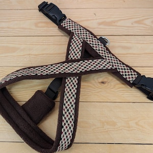 May include: Brown dog harness with a checkered pattern in cream, black, and red. The harness has a soft, padded interior and adjustable black plastic buckles for a secure fit. The design is laid out on a light wooden surface.