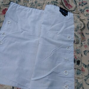 May include: Close-up of a white fabric curtain liner with white grommets along the edge. The liner is attached to a beige fabric curtain with a floral pattern.