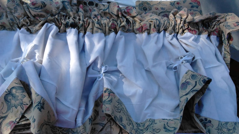 May include: A white sheer curtain with a floral patterned border. The curtain has a gathered look and is tied back with white ribbon.