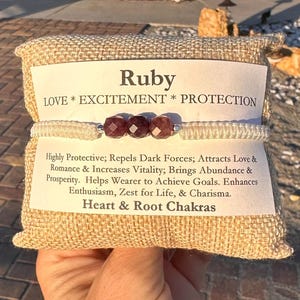 May include: A ruby bracelet with three faceted red gemstones, strung on a white cord. The bracelet is displayed on a burlap pouch with a label that reads "Ruby LOVE * EXCITEMENT * PROTECTION". The label also lists the benefits of the bracelet.