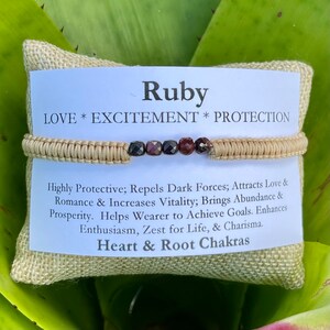 May include: A tan and red beaded bracelet with the words "Ruby, LOVE * EXCITEMENT * PROTECTION, Highly Protective; Repels Dark Forces; Attracts Love & Romance & Increases Vitality; Brings Abundance & Prosperity. Helps Wearer to Achieve Goals. Enhances Enthusiasm, Zest for Life, & Charisma. Heart & Root Chakras" on a tan burlap background.