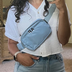 May include: A woman wearing a white shirt with a blue fanny pack that says "A.P." on it. She is wearing light blue jeans and a watch.