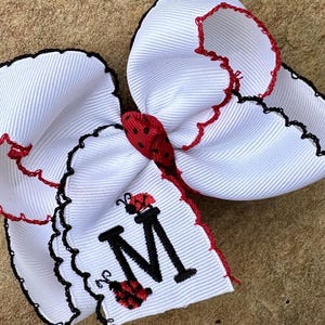 May include: White hair bow with red and black accents. The bow features a ladybug design and the letter "M" embroidered in black. The edges are trimmed with black and red stitching, creating a decorative border.