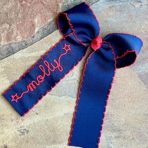 May include: A navy blue hair bow with red trim and a red center knot. The bow features the name "molly" embroidered in red with a star accent. The bow is set against a stone background.