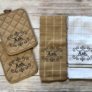 May include: A set of kitchen towels and pot holders with the name "Ash" embroidered on them. The towels are brown and white with a grid pattern. The pot holders are brown with a quilted diamond pattern. One pot holder has the letter "A" embroidered on it.