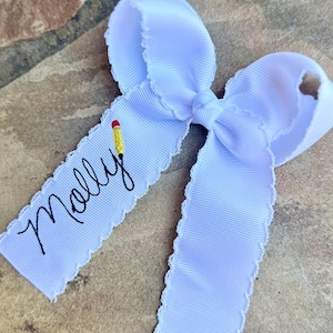 May include: A white ribbon bow with a scalloped edge. The bow has a tag with the name "Molly" in black script and a small pencil illustration. The bow is on a textured, brown surface.