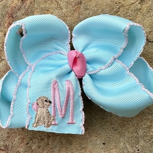 May include: A light blue hair bow with a white scalloped edge. The bow has a pink center and a brown and gold embroidered dog with the letter "M" in pink.