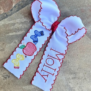 May include: White hair bow with red trim and a red center. One side features an embroidered apple, pencil, and socks. The other side has the name "Molly" embroidered in red.