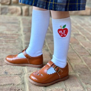 May include: White knee-high socks with a red apple embroidered on one sock. The apple has a green stem and leaf and the letter "M" in the center. The socks are worn with brown leather shoes with a buckle.