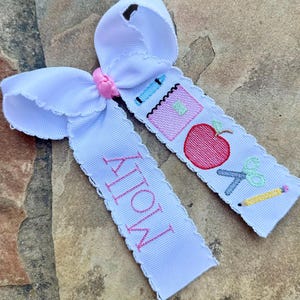 May include: White hair bow with a pink center knot and two ribbon tails. One tail has the name "Molly" embroidered in pink. The other tail features embroidered school-themed images: a crayon, notebook, apple, scissors, and pencil.