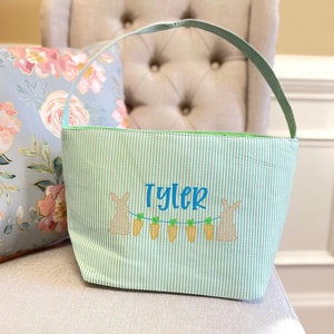 May include: A green and white striped Easter basket with a personalized name "Tyler" embroidered on the front. Two bunnies are embroidered on either side of the name with carrots hanging between them.