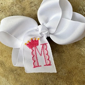May include: White bow with a pink letter M and a gold crown on a white tag. The letter M is surrounded by a pink scalloped border.